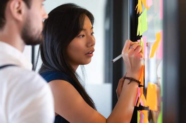 A woman in an office writing on sticky notes on a window while a man stands beside her.