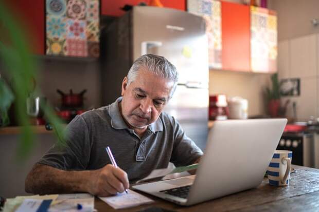 Person sitting at kitchen table in front of laptop writing note.