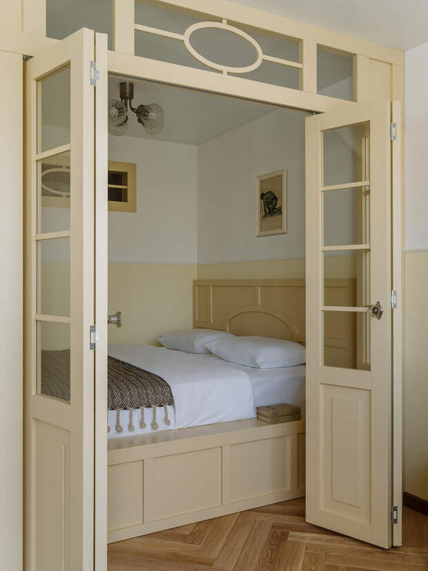 Большую кровать разместили в специально оборудованной нише за распашными дверьми со стеклянными вставками (за счет этого в импровизированную спальню попадает солнечный свет)
