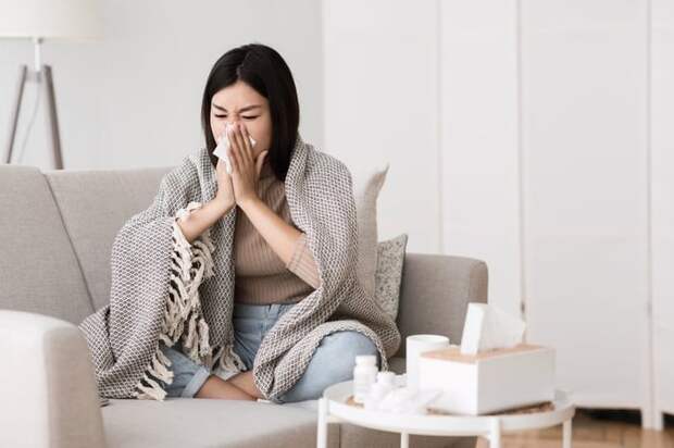 A woman sitting on a couch under a blanket and blowing her nose.