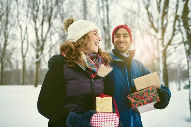 A smiling man and woman walking through a snowy park while holding wrapped gifts.