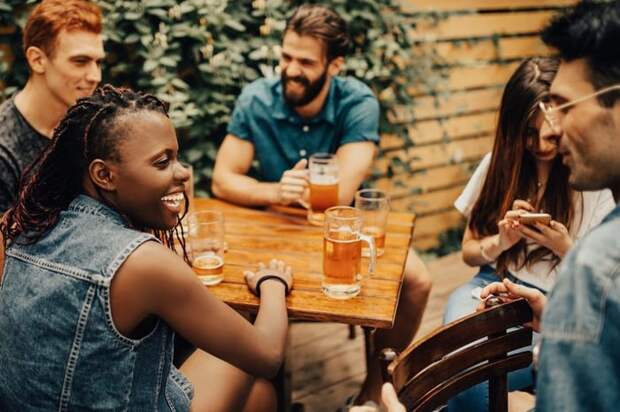 A group of laughing friends drinking beers at a restaurant patio table.