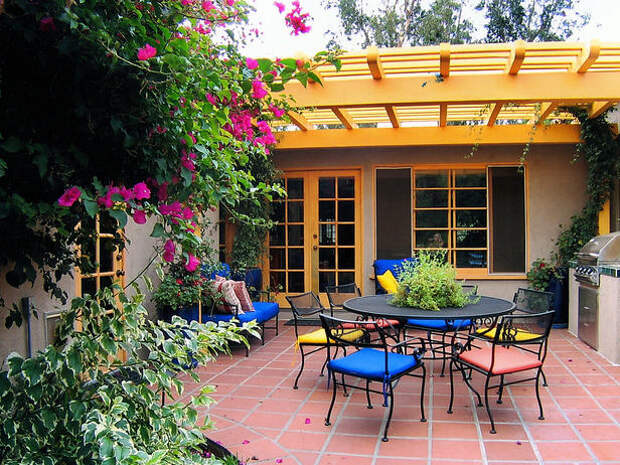 Wide view of shaded porch with black iron outdoor furniture, red tiled floor, outdoor grill, plants and flowers.