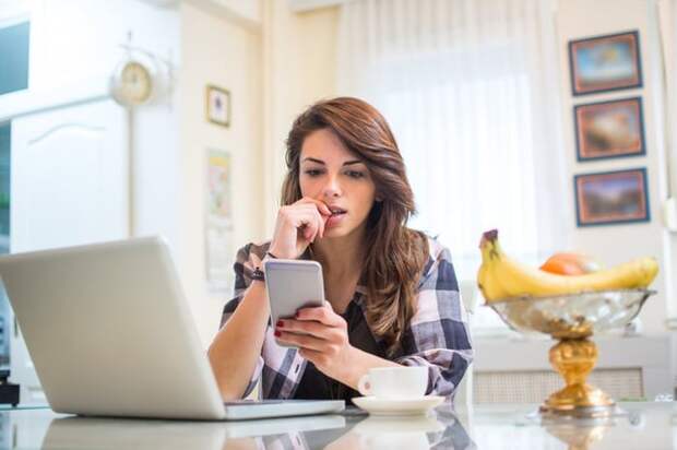 A nervous young woman biting her fingernail while sitting in her kitchen with her phone and laptop.