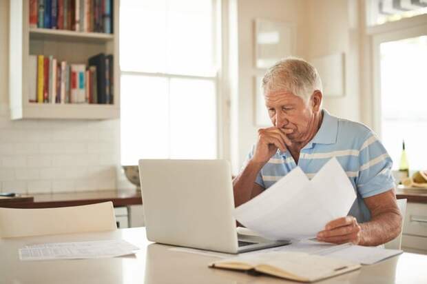 A stressed person looking through papers and sitting in front of a laptop at a kitchen counter.