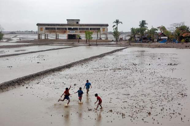 Неунывающий дух.  Дети играют в Джалиахали,- село, которое было полностью разрушено циклоном Айла в 2009 году. Этот шторм заставил жителей состязаться за один из нескольких сотен приютов от циклона (на фото сверху), большинство которых функционируют как центры для общин.