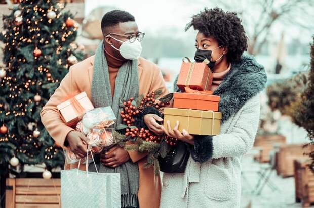 Two people wearing masks and carrying wrapped gifts while walking down a street decorated for the holidays.
