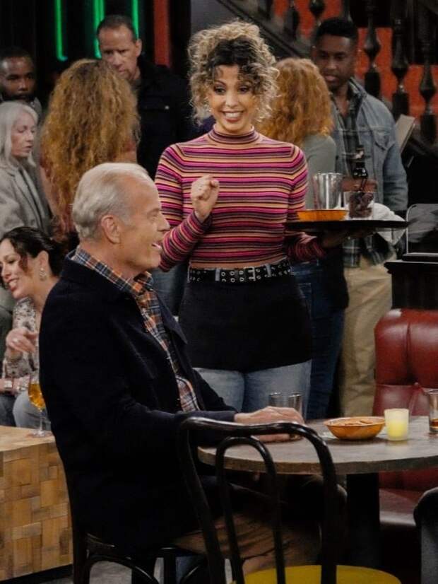 Frasier sits and Eve stands with a tray of drinks at the local bar