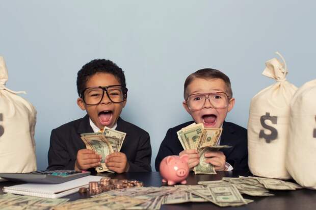 Two young boys wearing glasses and business suits holding money in their hands while smiling, and sitting next to several money bags.