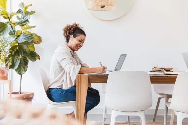 A smiling person sitting at their dining table and writing in a notebook next to an open laptop.