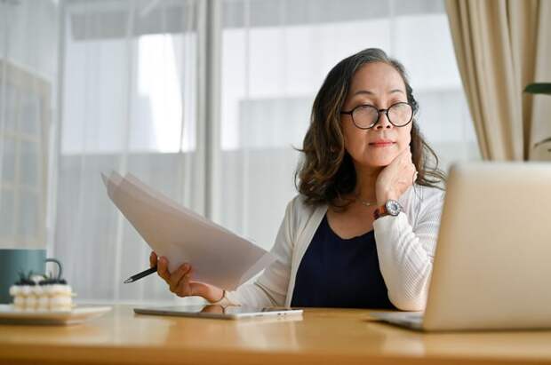 A businessperson holding paperwork in their right hand while looking at an open laptop on their desk.