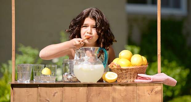 The sun shines on a girl tasting lemonade at her outdoor lemonade stand. 
