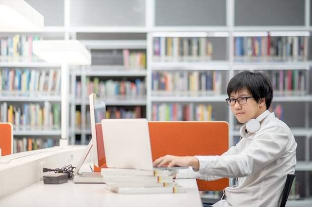 A male college student in the library with his laptop and books.