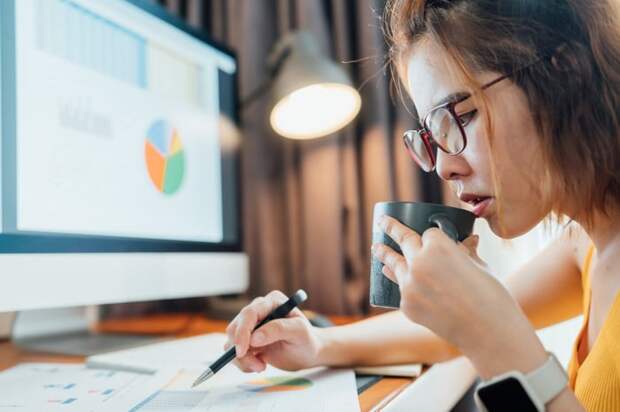 Investor at a desk sipping from a mug and looking at financial charts on papers and a computer monitor.