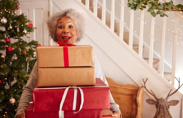 A woman standing in her decorated living room holding an armful of gifts.