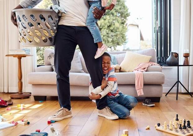 A parent holding a laundry basket in one arm and a child in the other while another child grabs their leg.