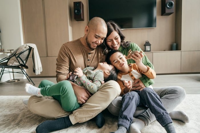 A smiling mom and dad sitting on the floor with their young kids playing in their laps.