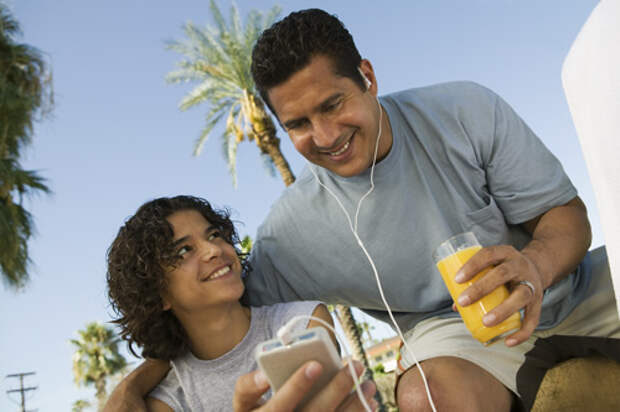 Boy (13-15) holding portable music player, father listening with earphones and holding glass of juice.