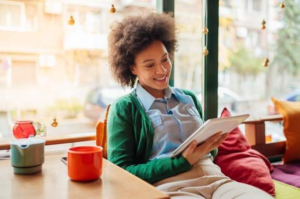 A woman sitting in a sunny window seat and checking her tablet.