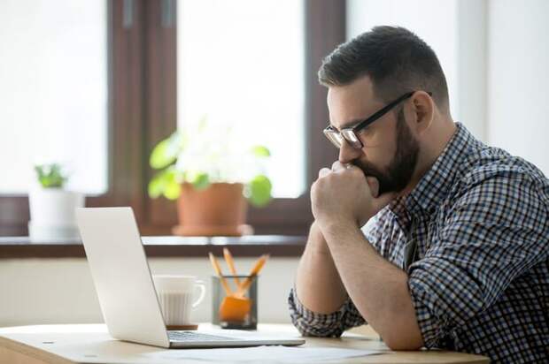 A stressed man sitting at a desk in front of an open laptop with his chin in his hands.