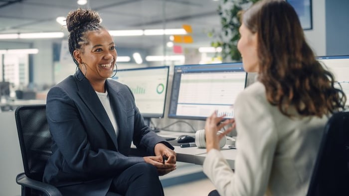 Two smiling people sitting at their office desks in front of their computers and talking.