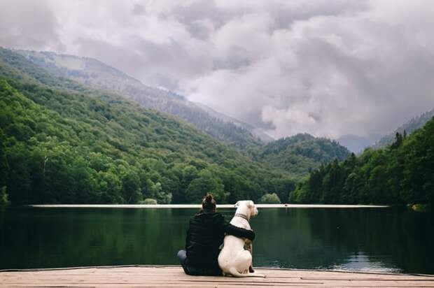 A woman with her arm around he dog sitting in front of a lake.