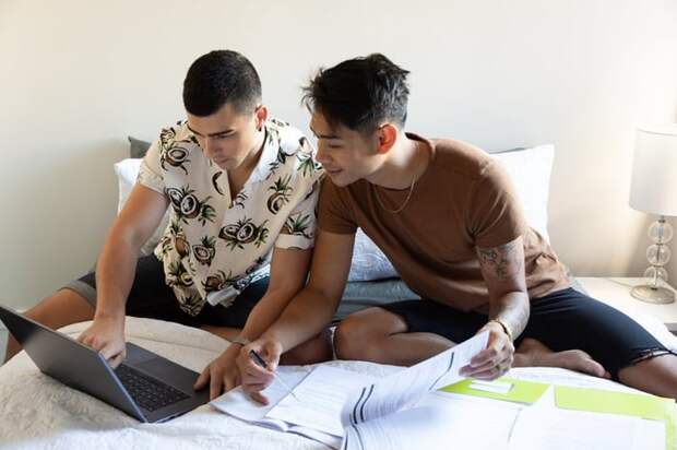 A couple sits on their bed looking at a laptop and paperwork.